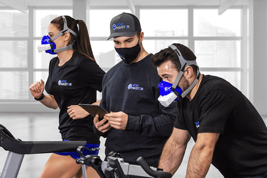 Athletes biking and running inside a gym, wearing the metabolic and performance analyzer while a coach reviews data on an iPad