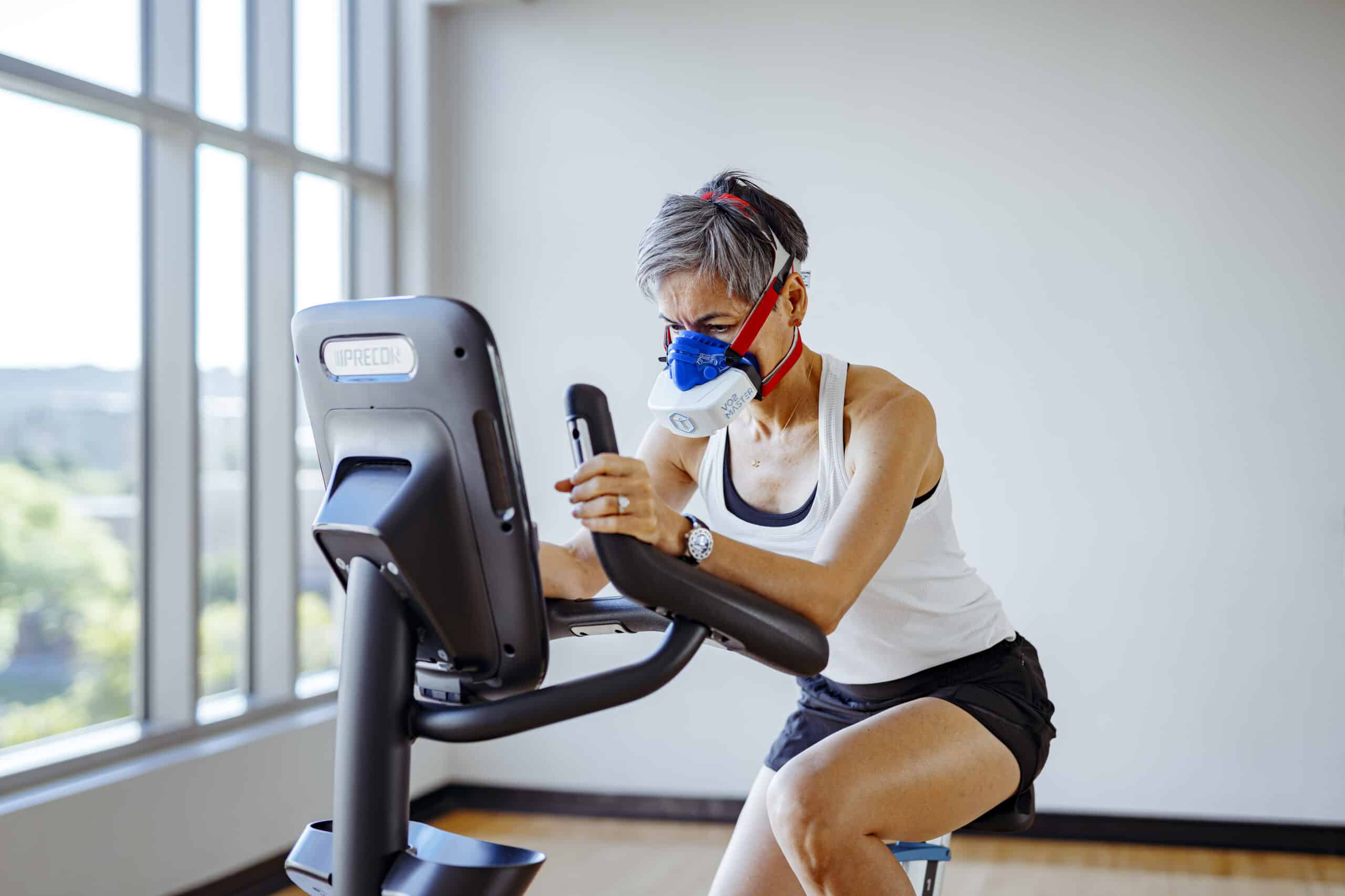 A middle-aged woman rides a stationary bike with the VO2 Master analyzer and a blue mask