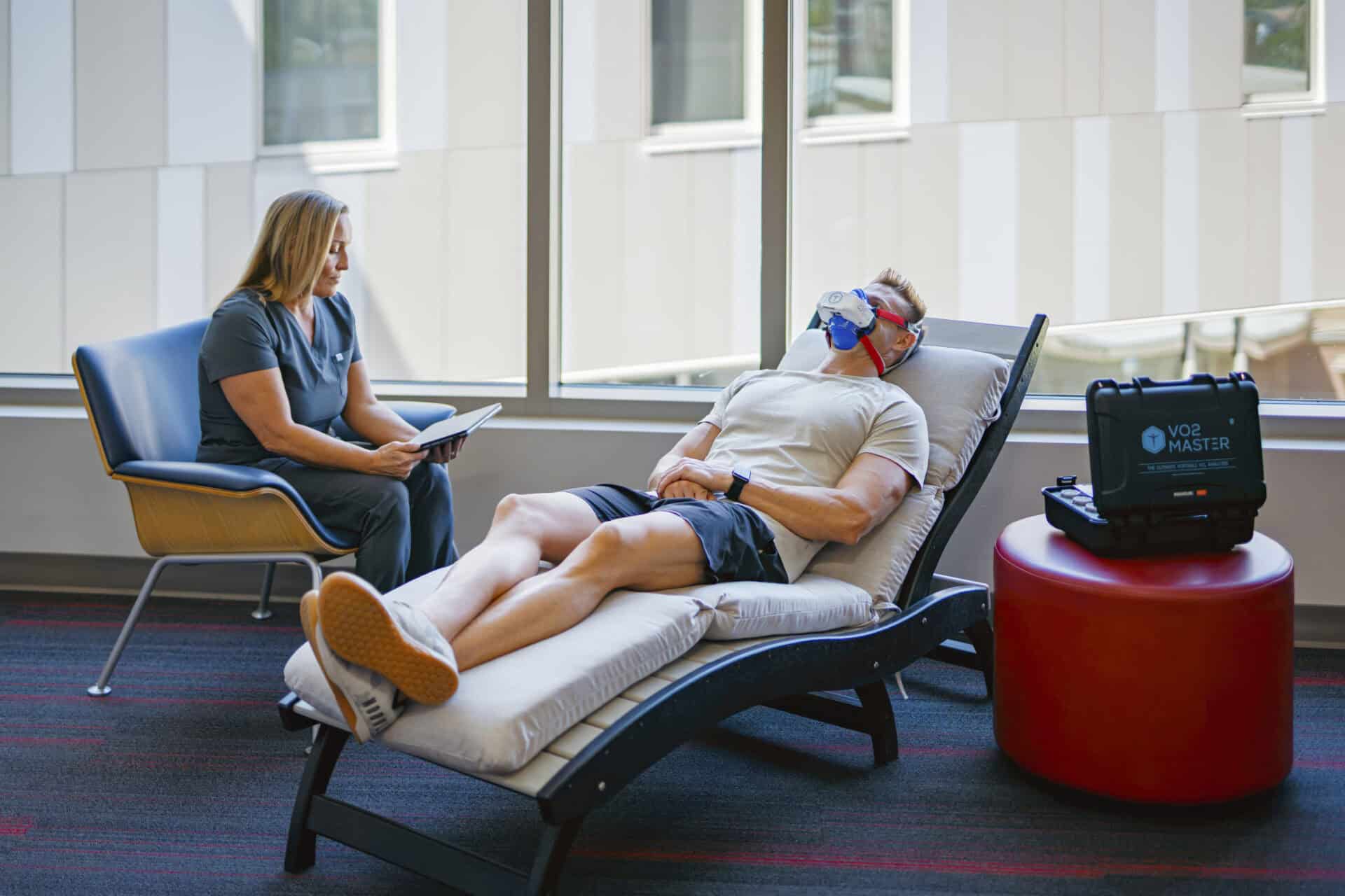 A fit middle-aged man lays on a daybed while a facility operator tests his recovery