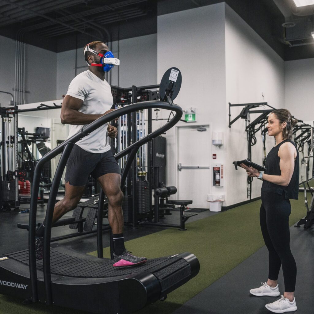 Male athlete running on treadmill inside a gym while female coach watches and measures via the testing app on an iPad