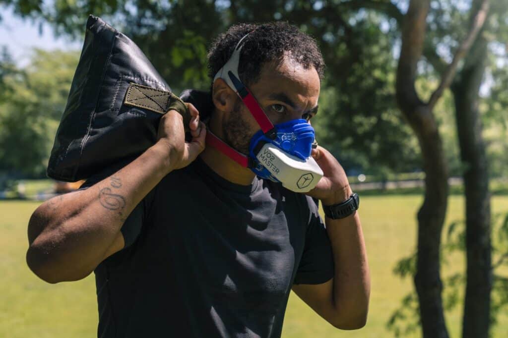 Man training for military, carrying sandbag over his head outdoors, wearing VO2 Master Analyzer and mask