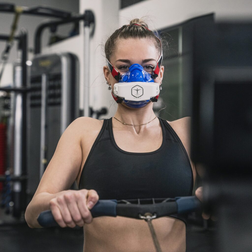 Woman rowing inside a fitness facility, wearing black sports bra and the VO2 Master analyzer and blue mask