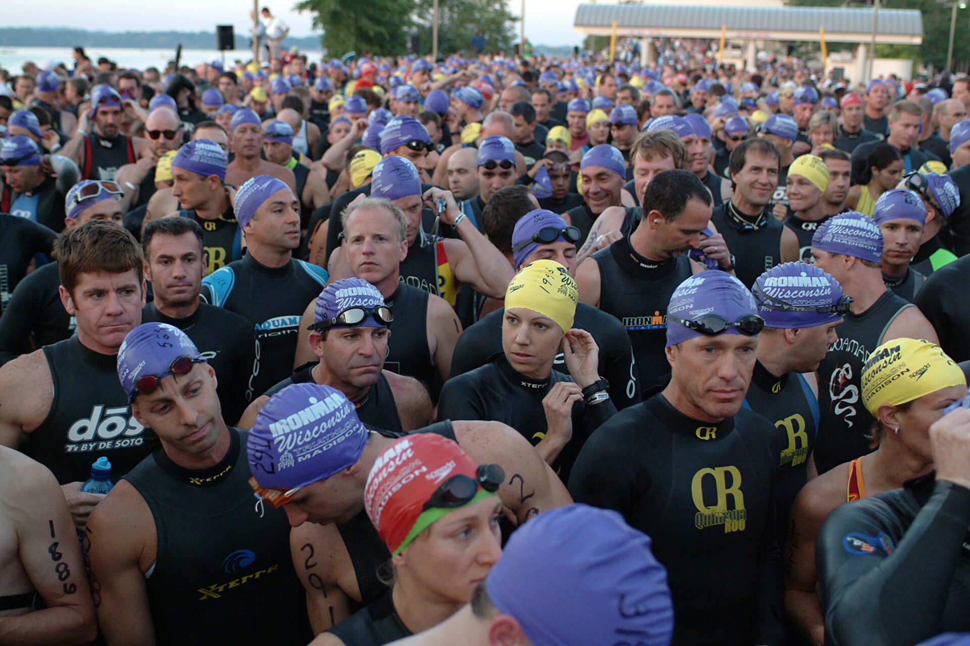 Triathletes are shown preparing for the swim portion of the triathlon in wisconsin.  They wear purple swim caps and stand together in a crowd ready to max out their training thresholds for the race.