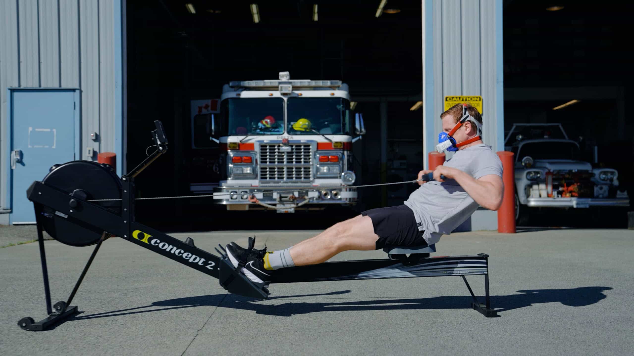 Firefighter performing metabolic test on rower using VO2 Master Analyzer outside fire station with fire truck in background.