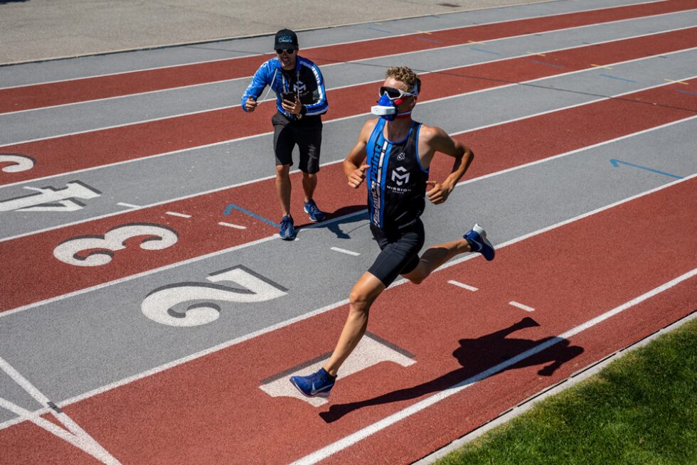 Runner sprinting on a track, measuring performance with a VO2 mask while trainer watches and records on iPhone