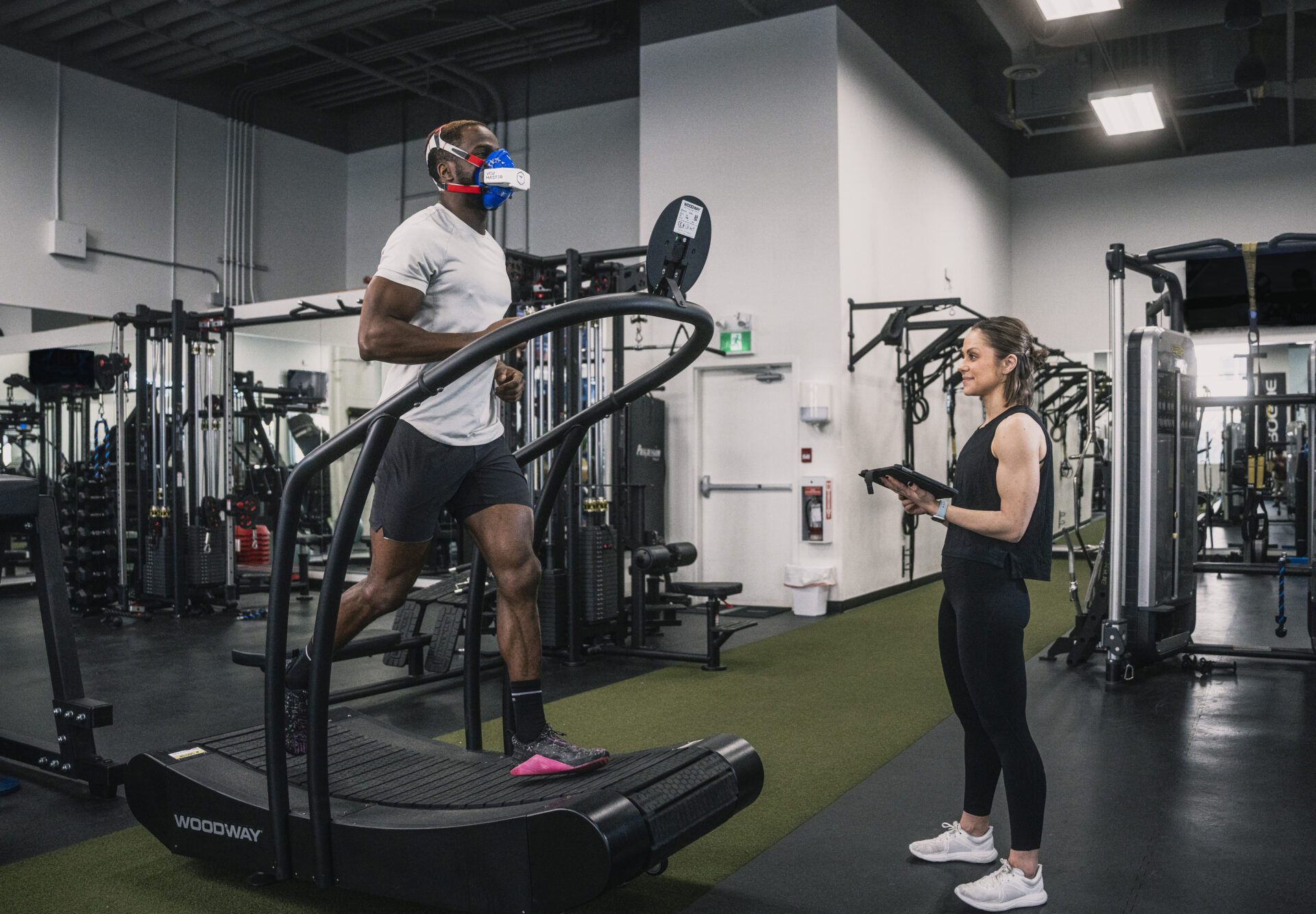 Male athlete runs on the treadmill while a female coach stands nearby holding an ipad to monitor the success of the vo2 master analyzer during various tests and training zones