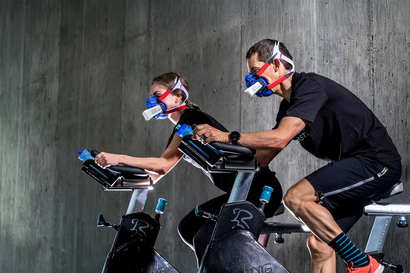 Two athletes performing VO2 max testing on indoor bikes using portable analyzers to track metabolic efficiency and longevity-focused performance metrics.