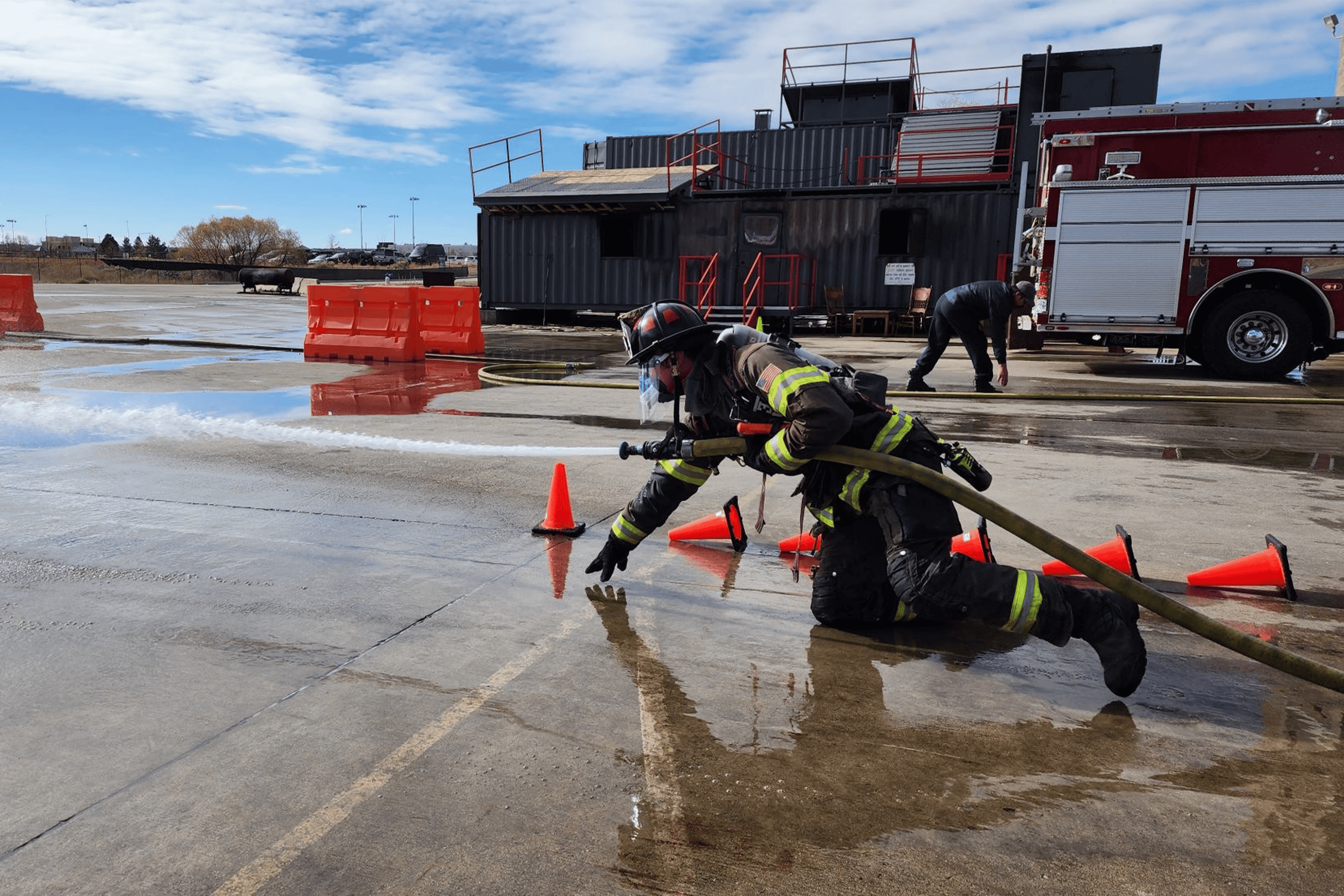 Firefighter wearing VO2 Master mask during live fire training drill, advancing hose through wet course with cones and training structures in background.