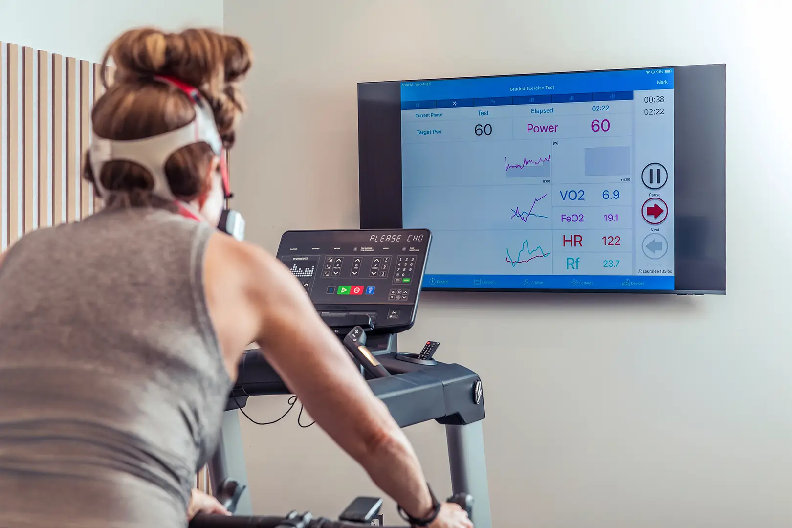 Benchmark Proactive female client completing a cycling test while live VO2, heart rate and respiratory data appear on a display.