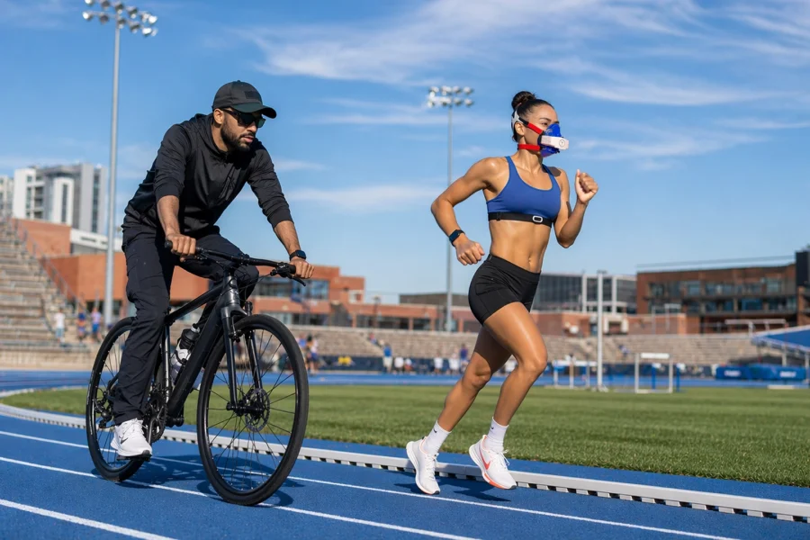 Female runner wearing a portable metabolic analyzer during track testing while a coach follows on a bicycle with phone showing live metabolic data via bluetooth.