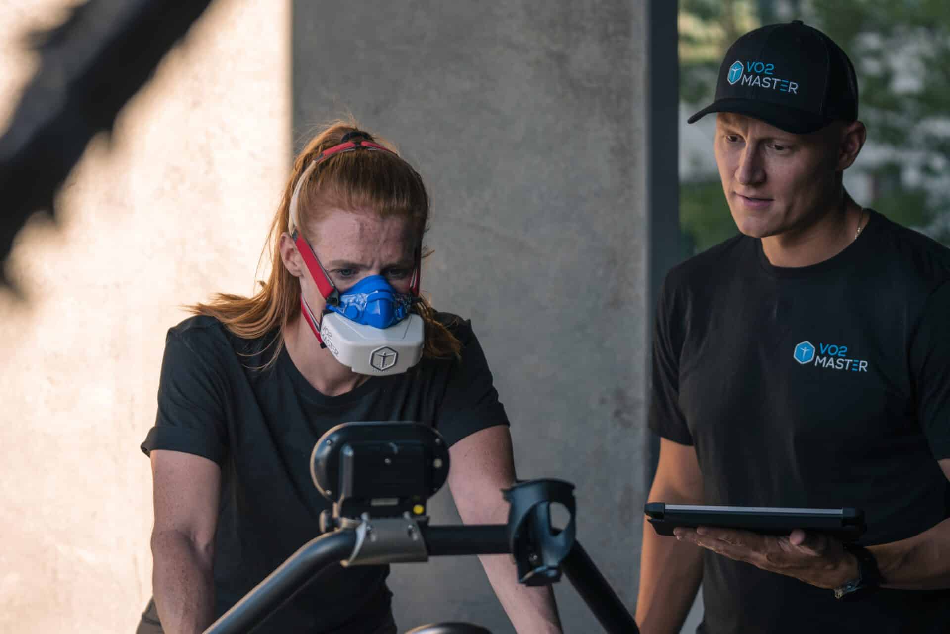 Woman biking in a fitness facility with a VO2 mask while trainer observes and monitors via an iPad