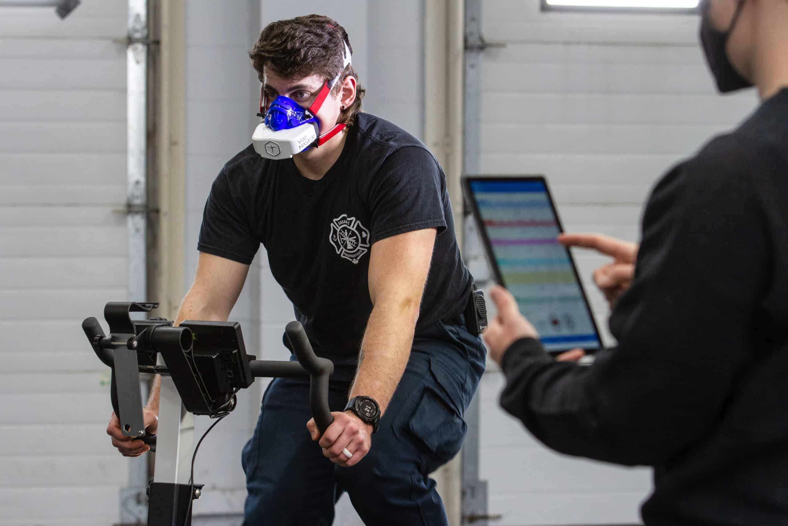 Firefighter wearing VO2 Master Analyzer conducting metabolic test on stationary bike while technician monitors live data on tablet.